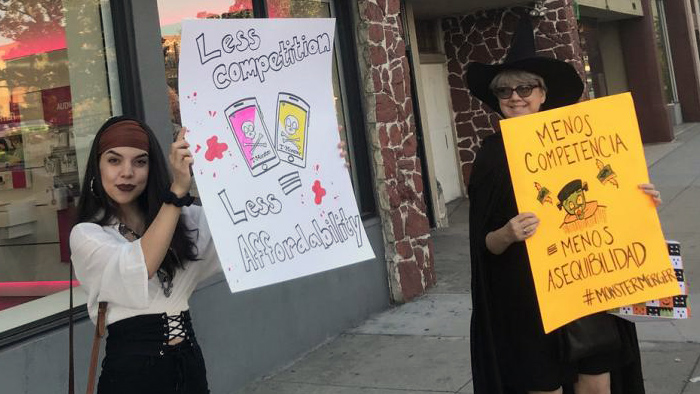 A woman dressed as a pirate and a woman dressed as a witch holding signs in English and Spanish reading “Less competition = less affordability”