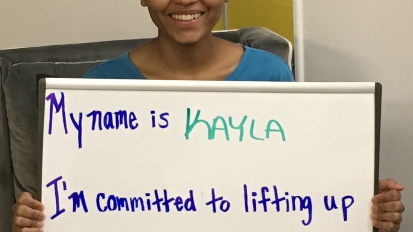 Woman holding a whiteboard saying "My name is Kayla. I'm committed to lifting up the stories of the first nation's people"