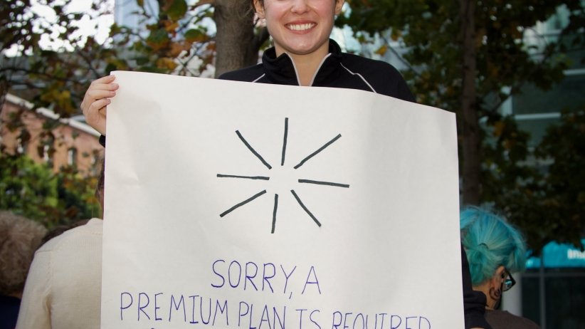 Activist holding sign reading "Sorry, a premium plan is required to view this poster"