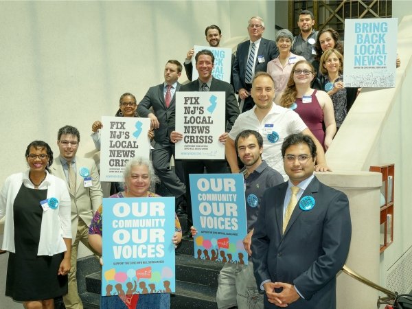 Group of people standing on stairs and holding signs reading "Our community / our voices" and "Fix NJ's local news crisis"