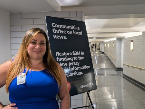 Free Press Action Journalism and Media Education Director Vanessa Maria Graber standing beside a sign in the New Jersey statehouse calling for more funding for the New Jersey Civic Information Consortium