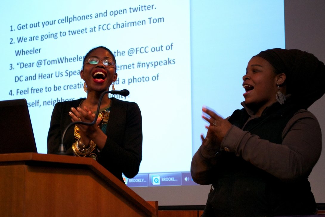 Two women at microphone in front of screen with text to Chairman Wheeler