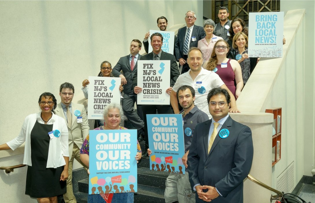 Group of people standing on stairs and holding signs reading "Our community / our voices" and "Fix NJ's local news crisis"