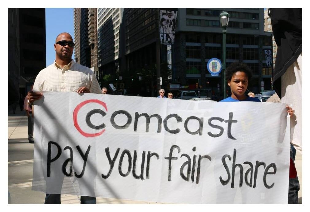 A father and son at a Philly protest holding a banner reading "Comcast: Pay your fair share"