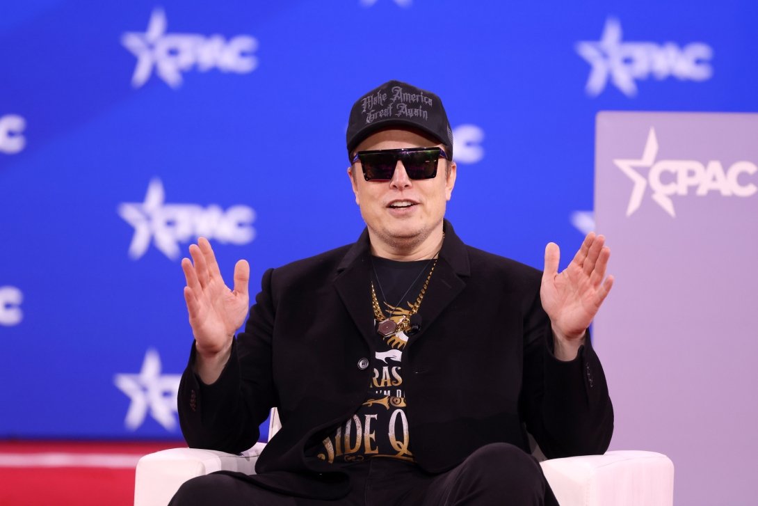 Elon Musk sitting and holding his hands up on stage at the 2025 Conservative Political Action Conference