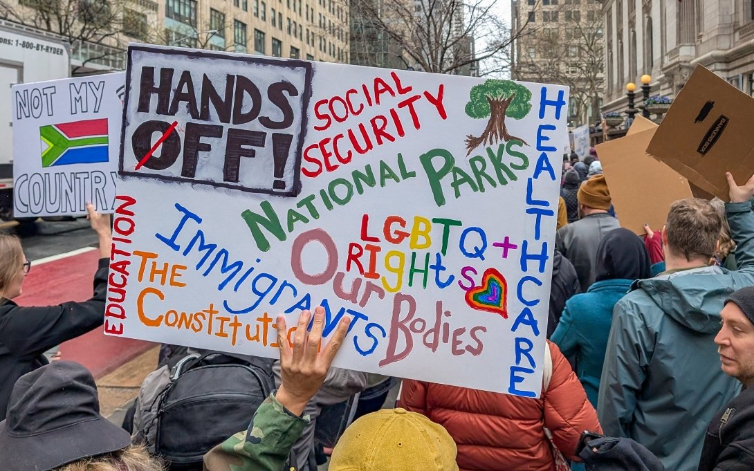 A photograph of a large group of protestors with one sign in focus. The sign in focus says Hands Off and lists a variety of issues.