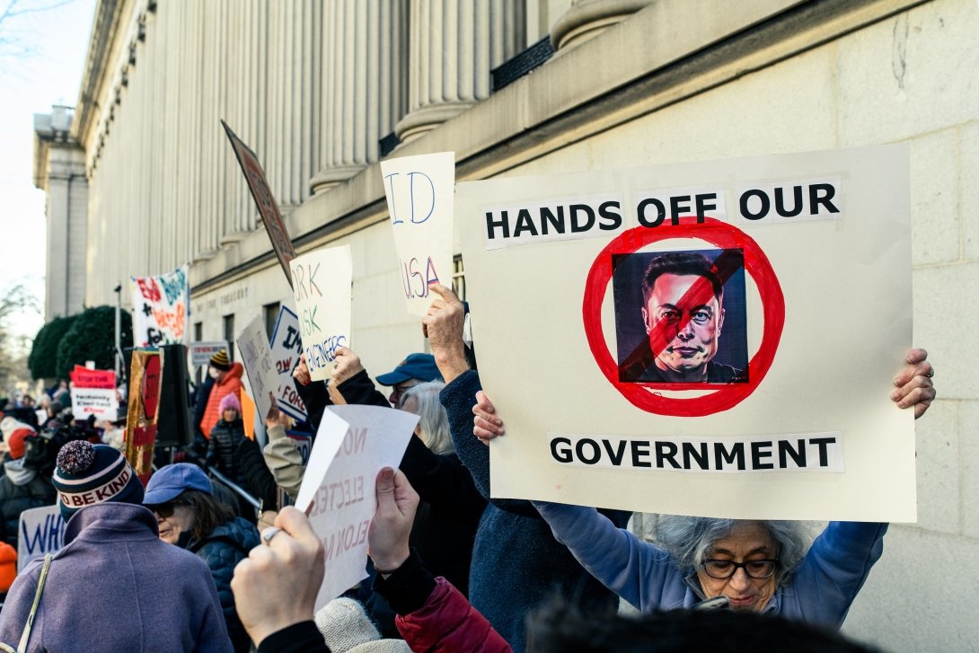 Protesters outside the U.S. Treasury Department holding a poster with an image of Elon Musk and the caption "Hands Off Our Government"
