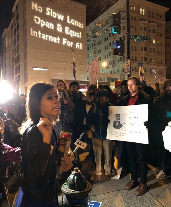 FCC Commissioner Jessica Rosenworcel speaking into microphone in front of activists; "No slow lanes / open and equal internet for all" is projected on building in the background