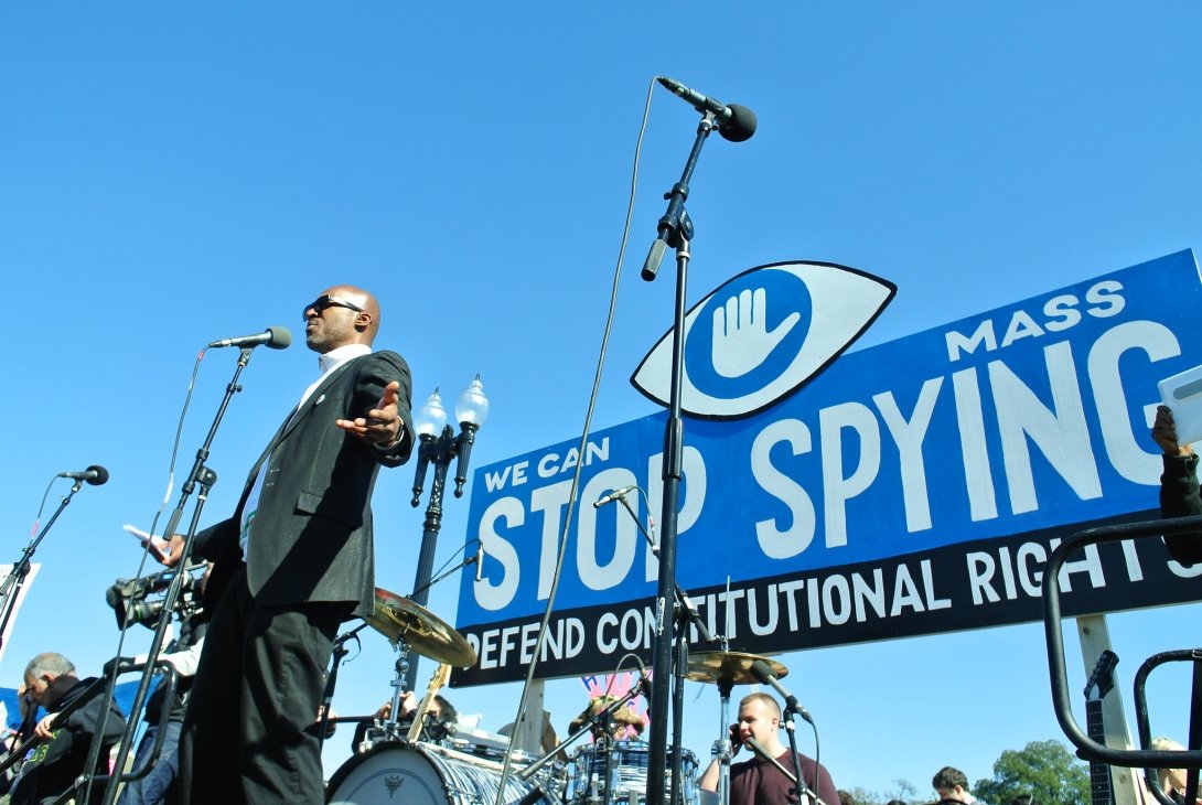 Man speaking at a protest before a banner reading "We can stop mass spying / defend constitutional rights"